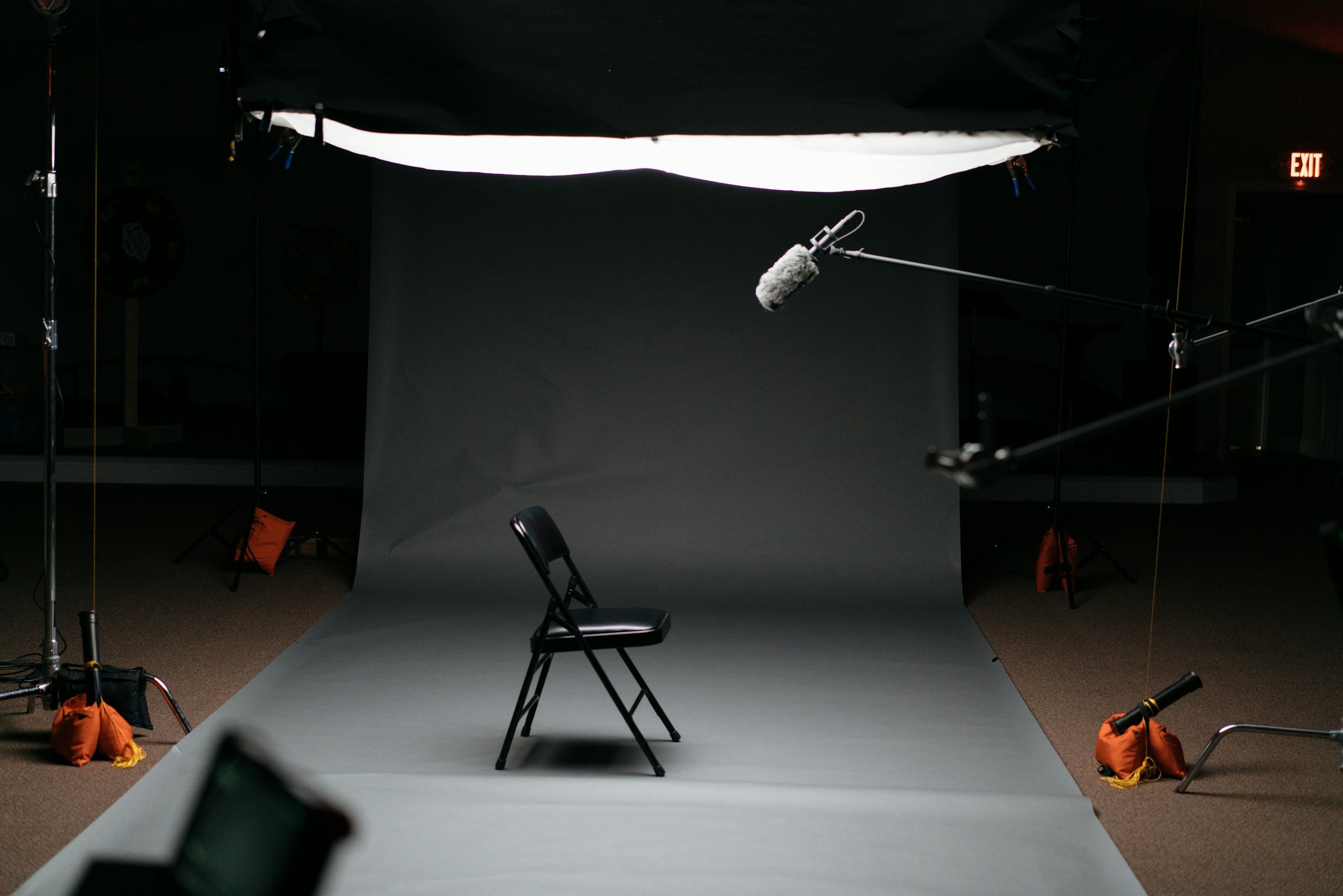 A photography studio setup with a gray backdrop, black chair, and various lighting and equipment items. A boom microphone with a fuzzy windscreen is positioned over the chair, and orange weights or sandbags are placed at the base of light stands and equipment supports to secure them.