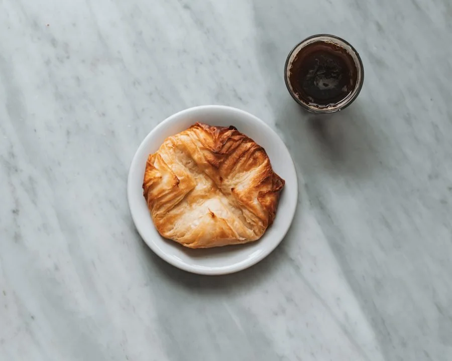 A croissant on a white plate with a glass of dark soda on a white marble surface.