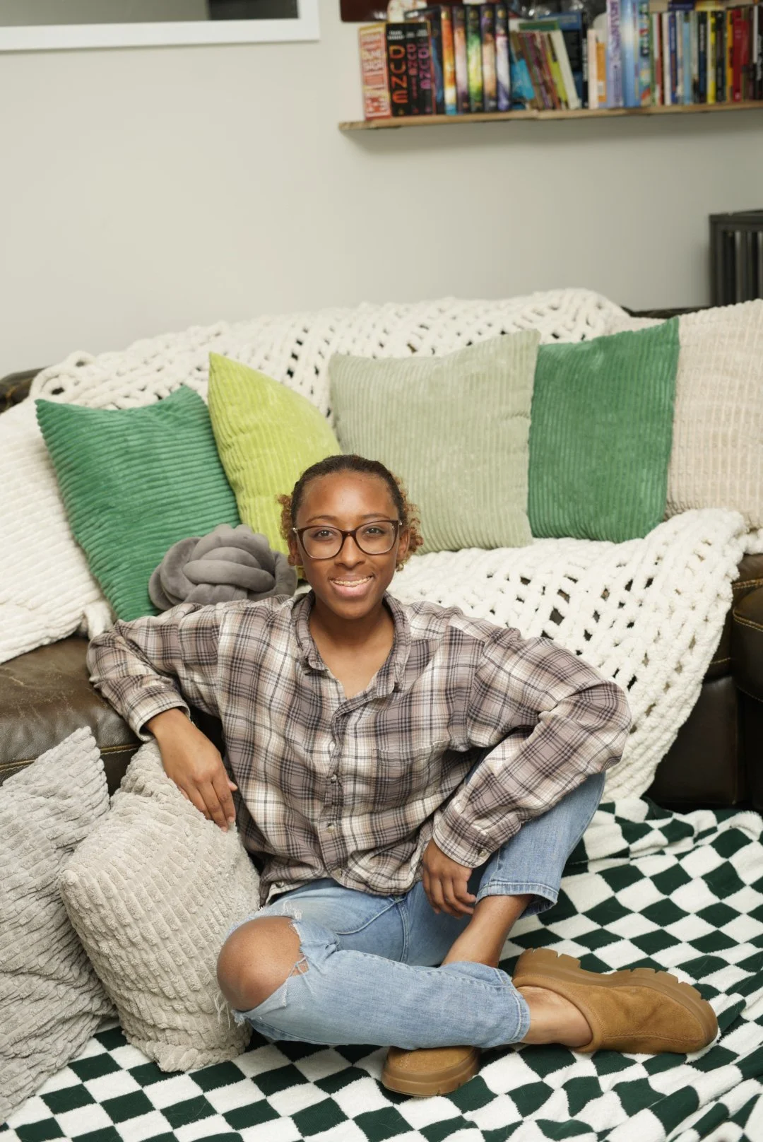 A woman sitting on the floor in front of a sofa with multiple colorful pillows and a blanket, in a room with a bookshelf on the wall.