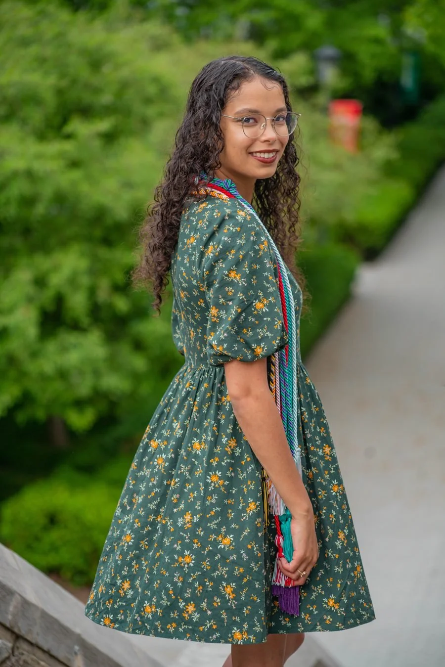 A young woman with long curly dark hair and glasses, wearing a floral dress, standing outdoors with a green, leafy background and a sidewalk.