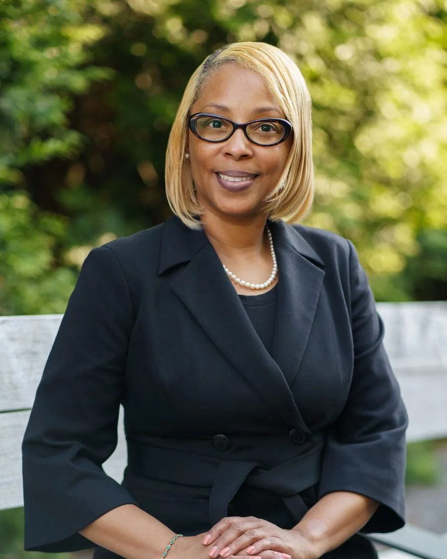 A woman in a black blazer and pearl necklace sitting on a bench outdoors with greenery in the background.