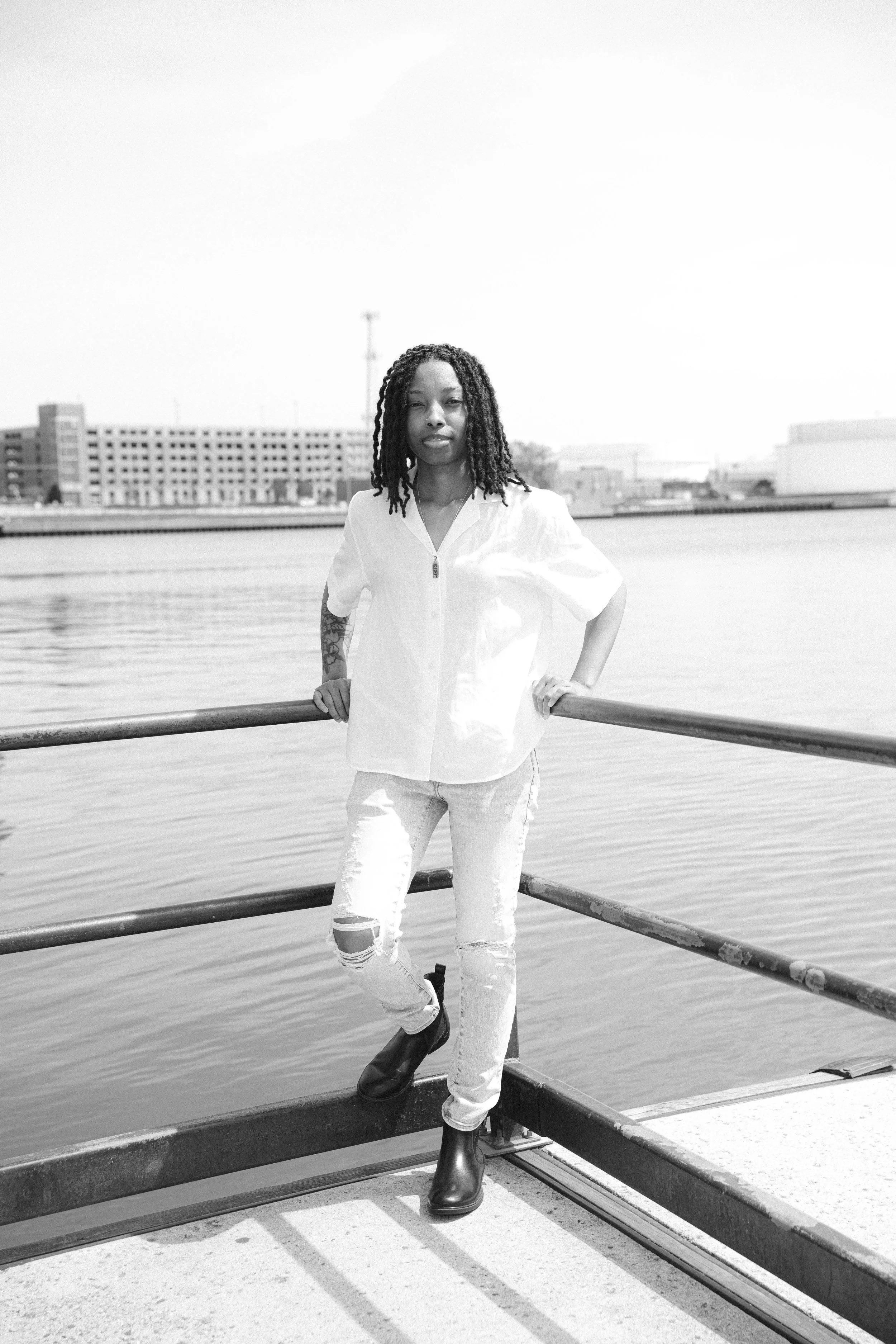 A woman with dreadlocks wearing a white zip-up shirt, ripped jeans, and black boots standing by a railing near water, with industrial buildings in the background.
