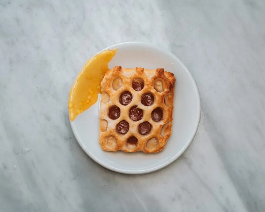 A white plate with a square honeycomb pastry topped with chocolate and a lemon wedge on the side, on a light gray surface.