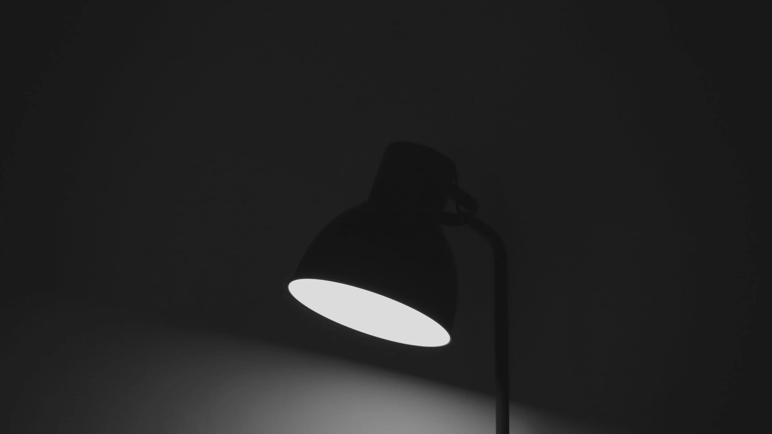 A black desk lamp with a white interior, turned on, casting light on a dark background.