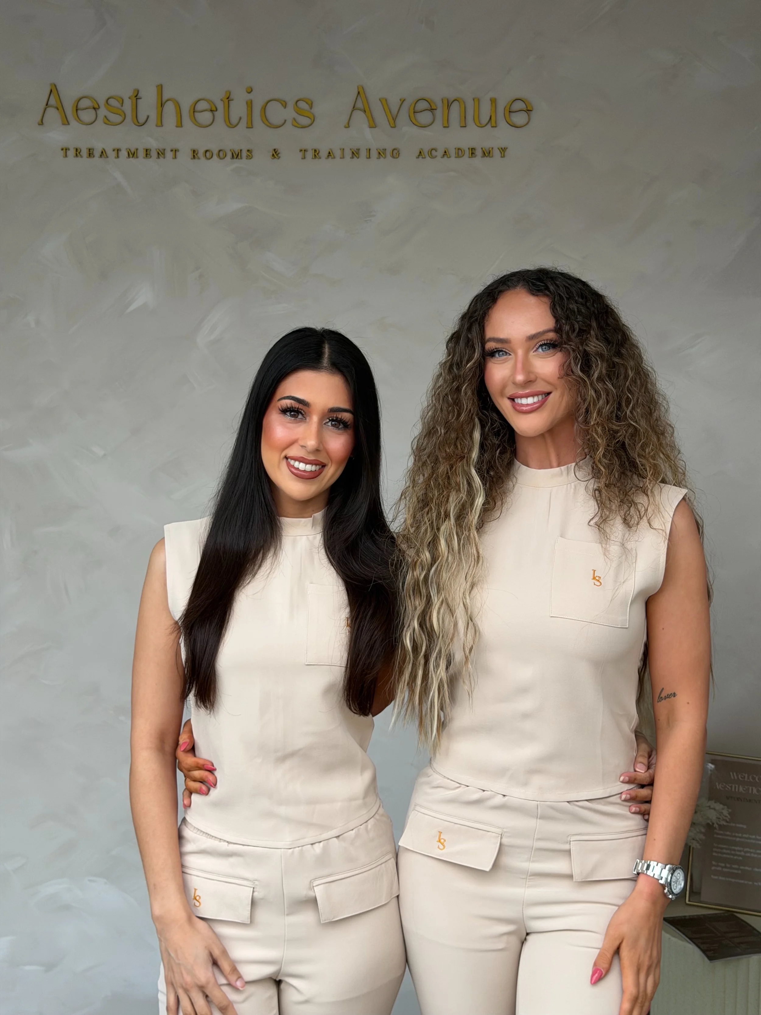 Two women standing side by side, smiling, in front of a sign that reads 'Aesthetics Avenue Treatment Rooms & Training Academy.' They are wearing matching beige outfits with small embroidered logos, and their arms are around each other's backs.