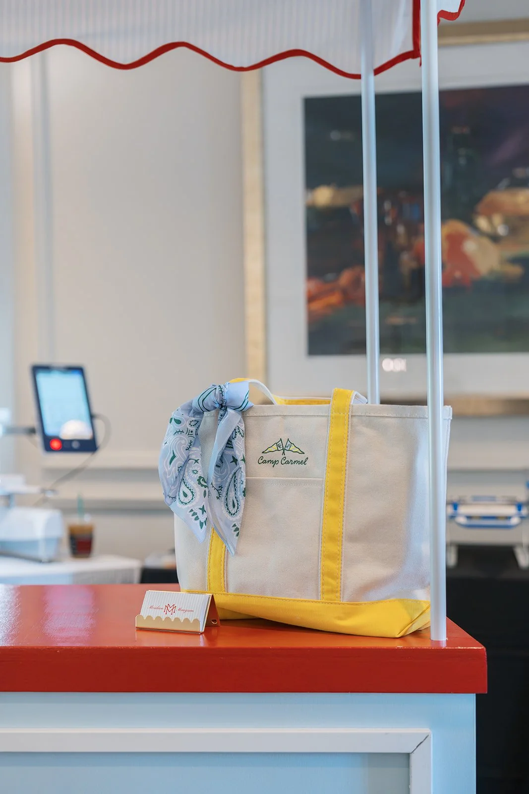 A white canvas bag with yellow trim labeled 'Camp Carmel' on a red countertop, with a blue and white scarf attached, in a reception area with a framed picture and a monitor in the background.