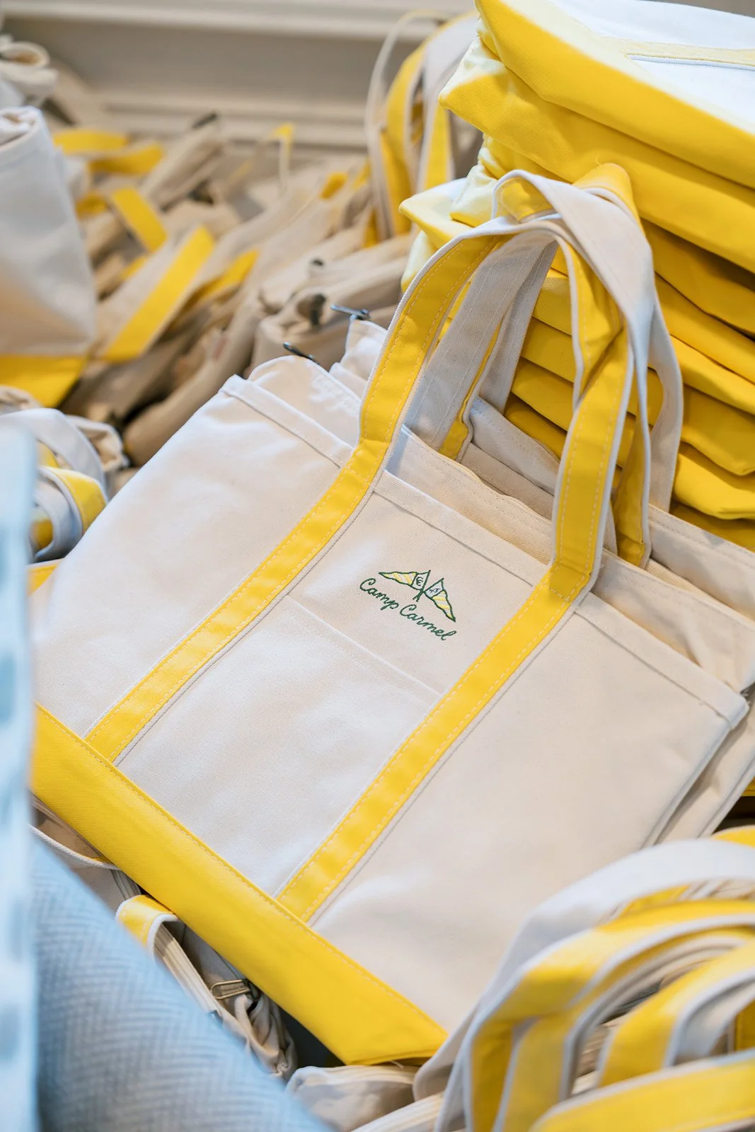 Stack of white tote bags with yellow trim, featuring a logo with a tent and the text 'Camp Carmel' at a store display.