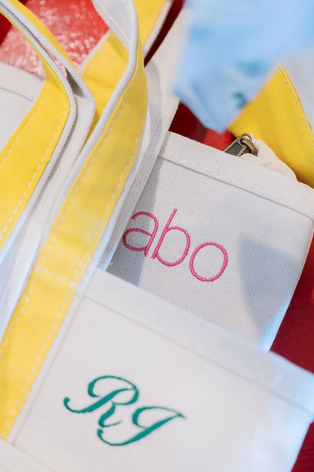 Close-up of personalized fabric tote bags with embroidered names 'abo' and 'Rqy', and a yellow and white bag with a zipper, on a red surface.