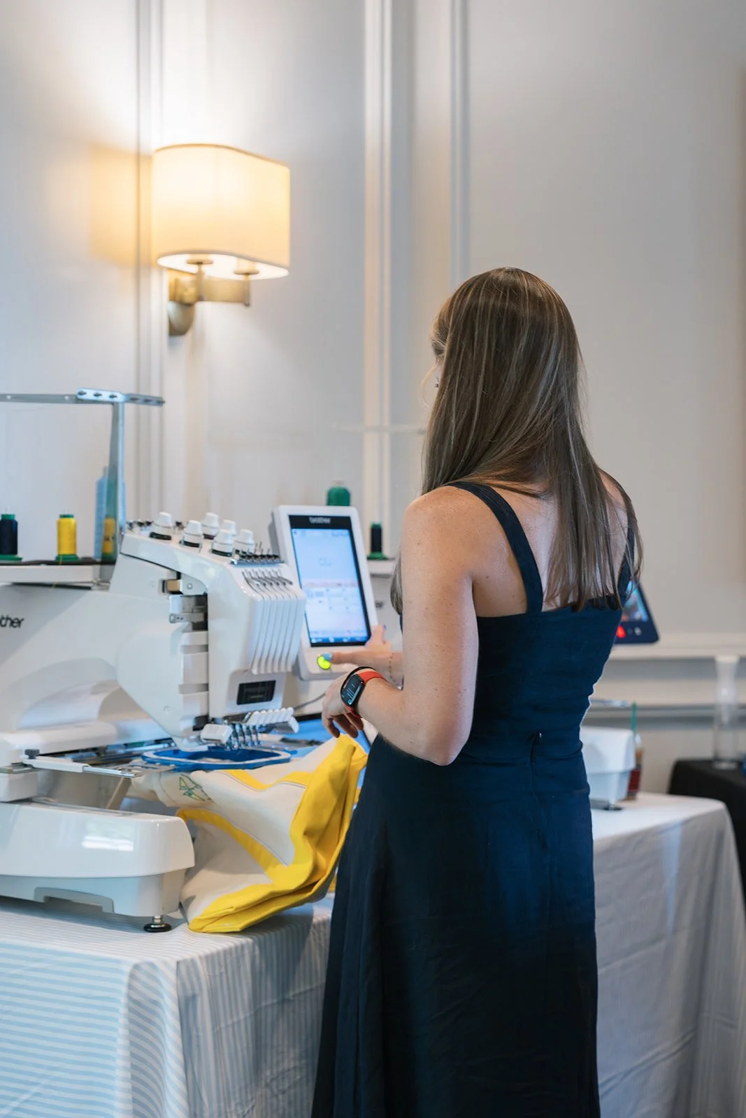 Woman operating embroidery machine at a table with fabric and supplies in a well-lit room.