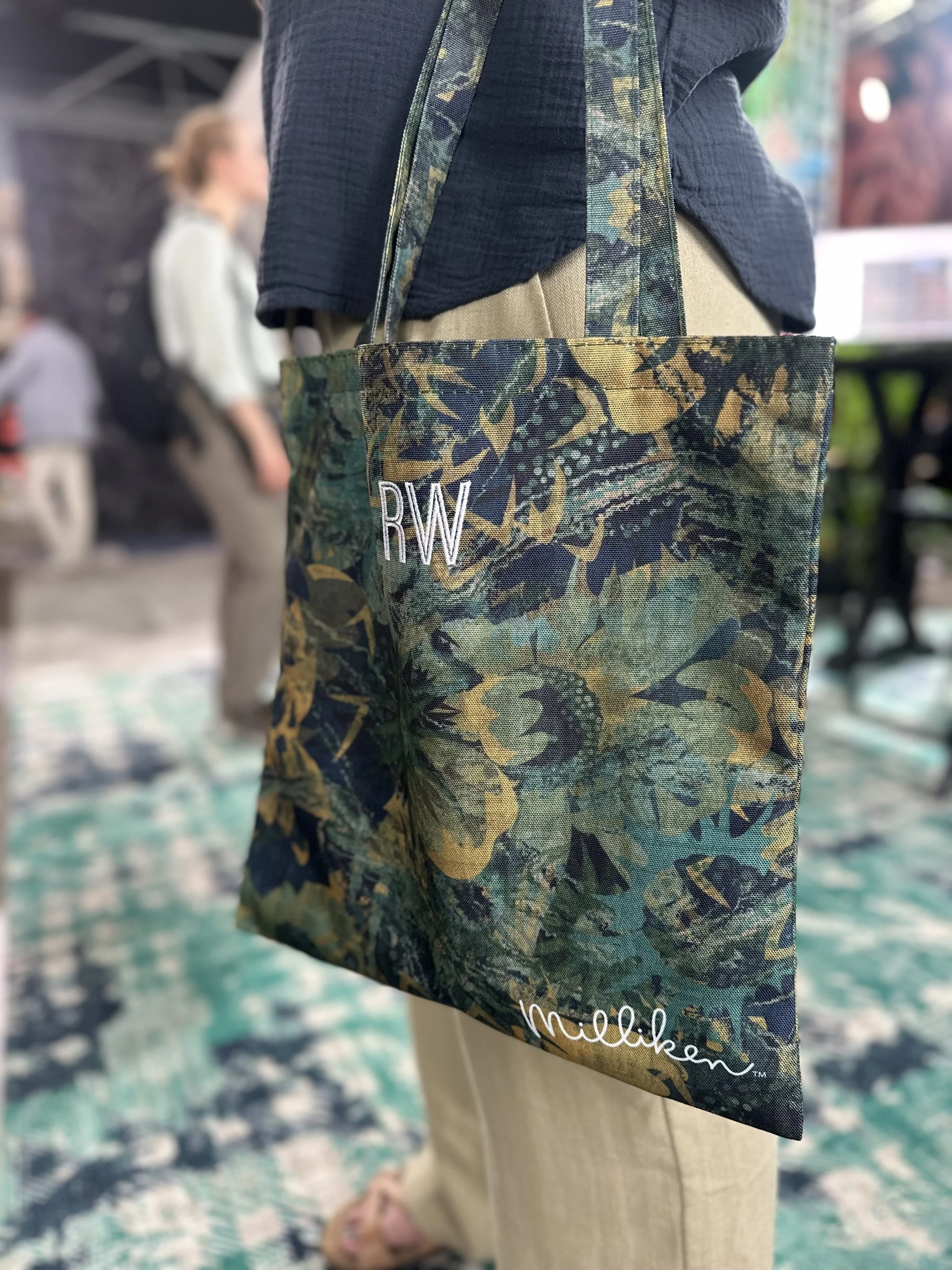 Close-up of a person holding a tote bag with a camouflage pattern. The bag has the initials 'RW' embroidered on it and the name 'Mille' written near the bottom. The person is wearing a dark top and beige pants, and the background shows other people and a colorful indoor setting.