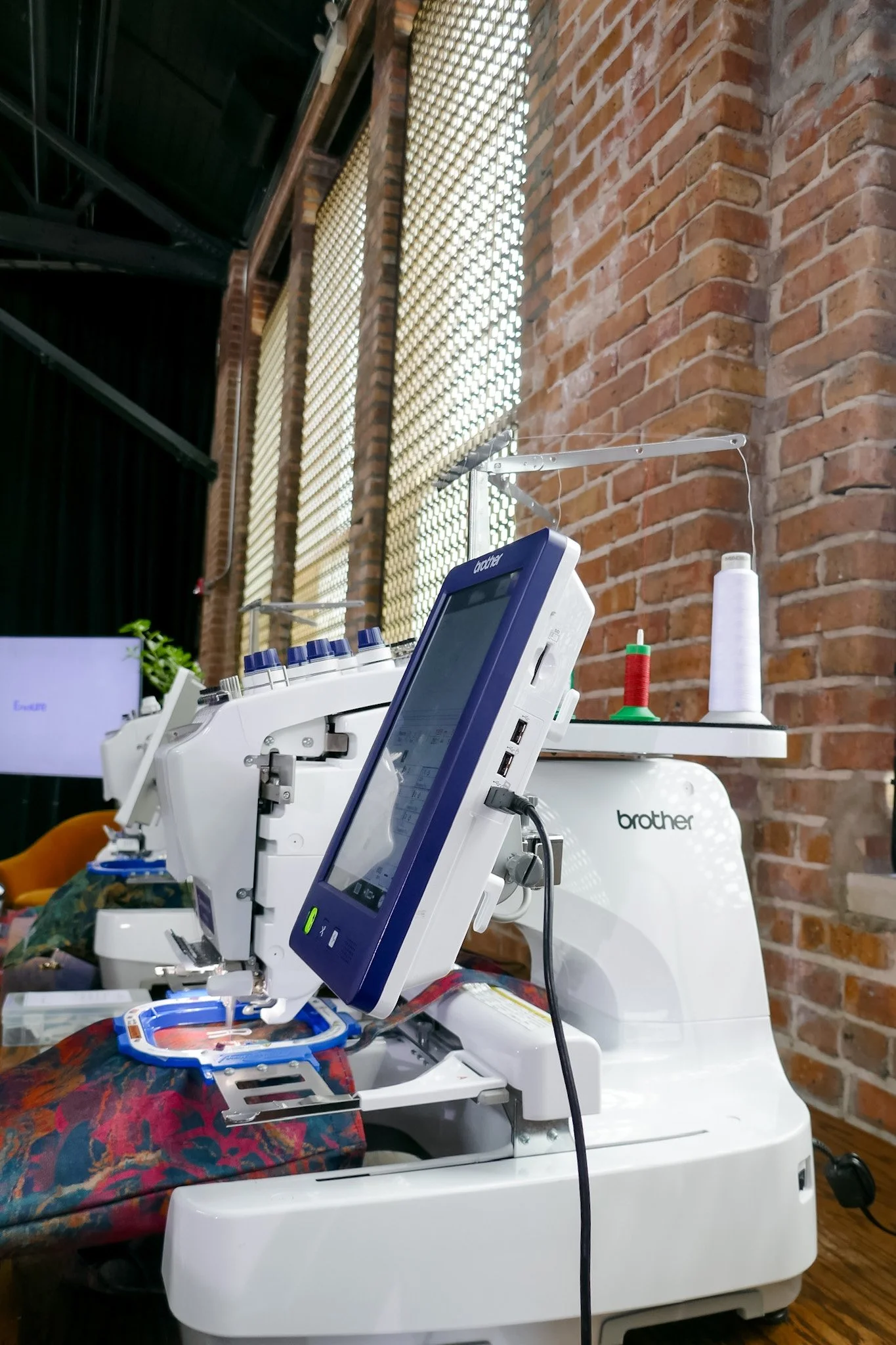 A sewing machine setup on a table in a room with brick walls. Spools of thread and sewing accessories are visible nearby. The room has large windows with wooden blinds.