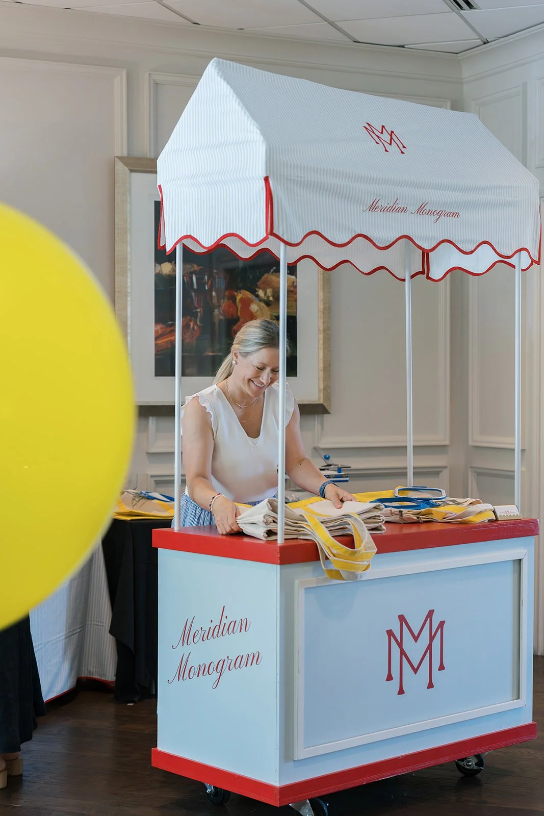 A smiling woman standing behind a merchandise cart with a white and red canopy, displaying tote bags with yellow straps, at an indoor event. The cart has the words "Meridian Monogram" and a logo.