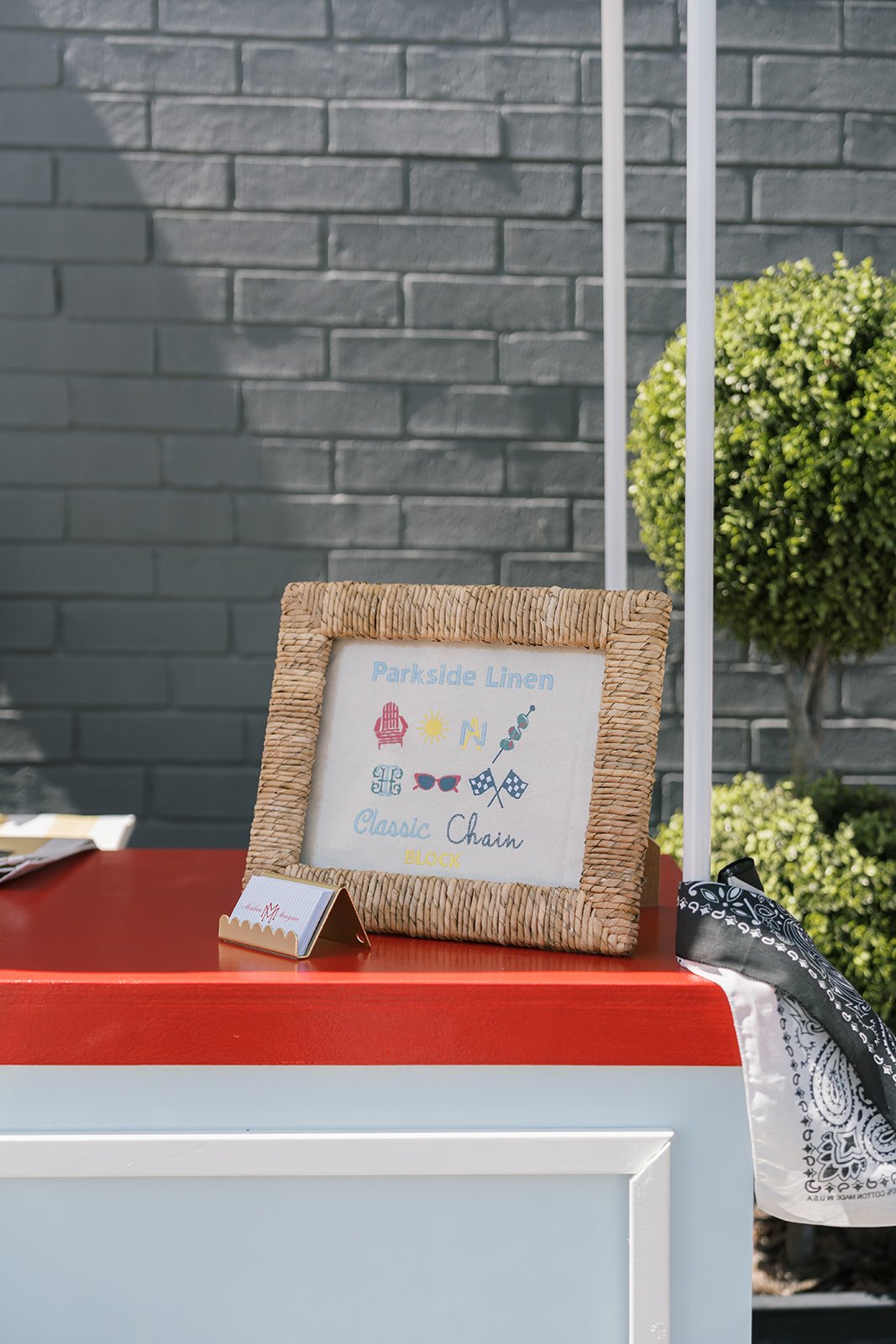A framed sign on a red and white counter reads "Parkside Linen" with summer-themed icons, including a beach chair, sun, and sunglasses, and phrases like "Classic Chain" and "Block"; a small folded paper sign is nearby, and a black and white bandana hangs over the counter's edge. In the background, there is a grey brick wall and a green potted plant.