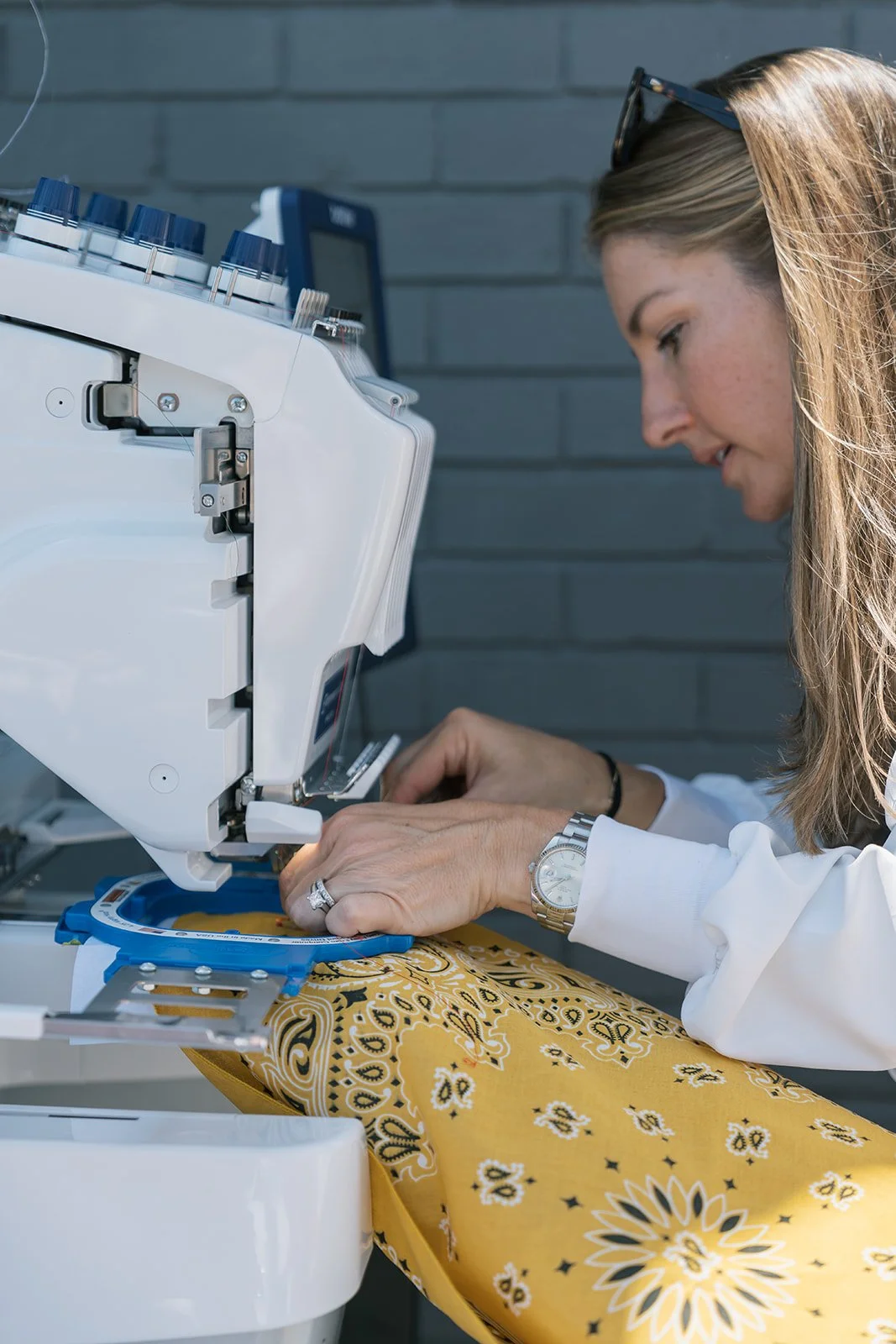 A woman sewing yellow bandana fabric with white and black floral pattern on an industrial sewing machine. She wears a silver watch and sunglasses on her head, with a grey brick wall in the background.