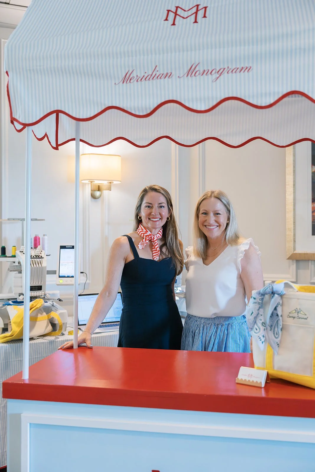 Two women standing behind a red and white booth at Meridian Monogram event, under a large umbrella with red piping and the Meridian Monogram logo, smiling at the camera.