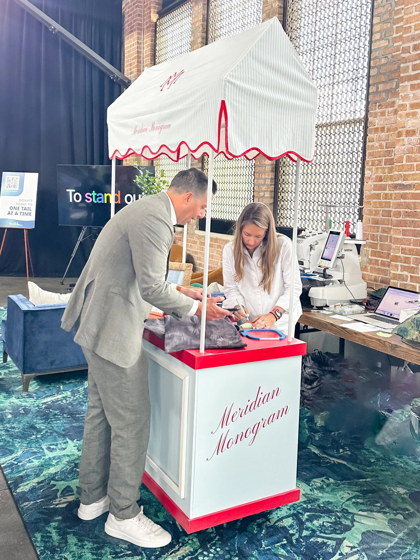 A man in a gray suit and white sneakers and a woman in a white shirt at a merchandise booth labeled 'Meridian Monogram' that is set up inside a building with exposed brick walls and large windows. There is a large screen behind them displaying the phrase 'To stand ou' with colorful letters. The booth has a white canopy with red trim and a red heart rate line design. There are laptops, printers, and other electronic equipment on the table.