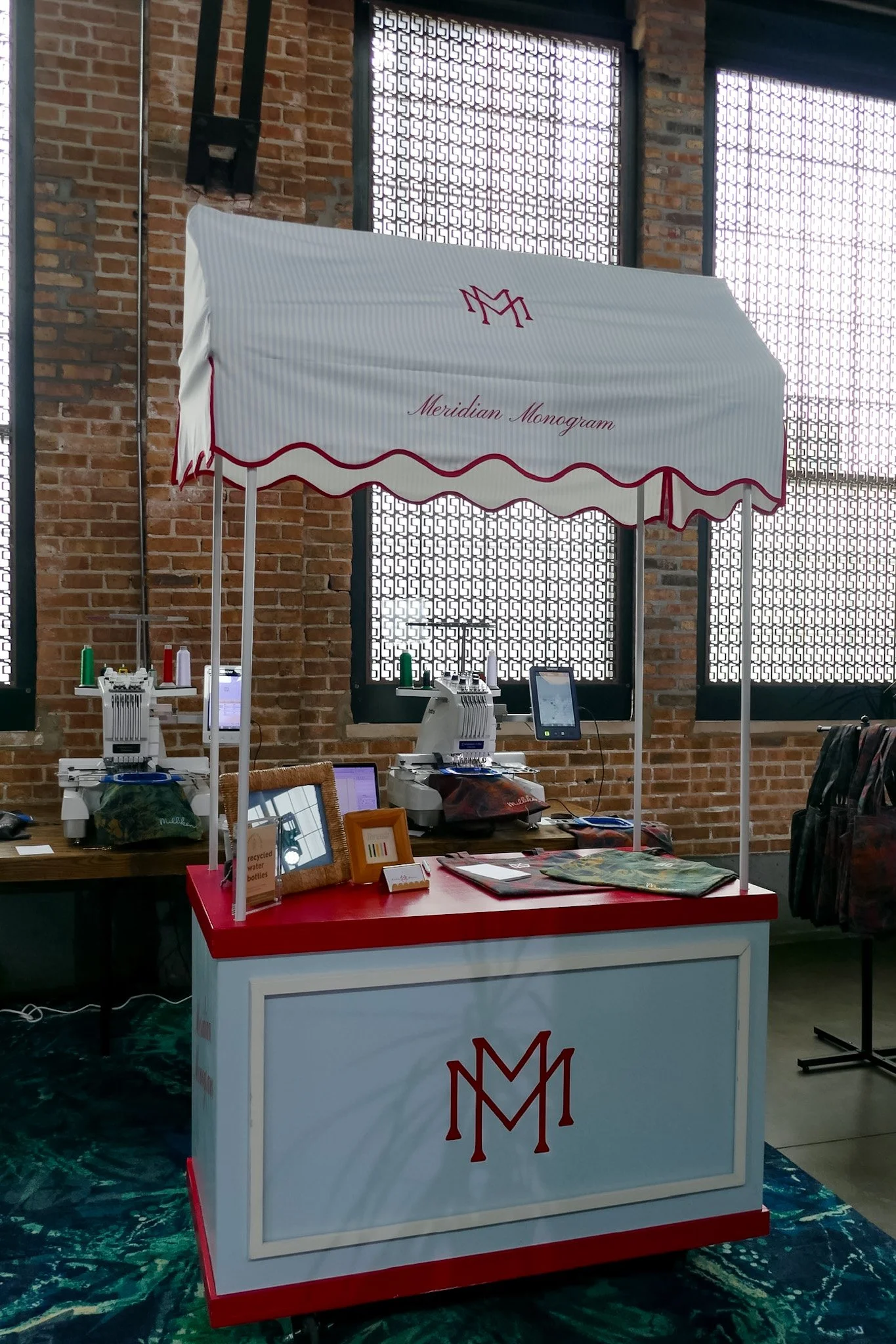 A trade show booth with a white canopy that reads 'Meridian Monogram' in cursive, supported by two poles, located inside a brick-walled building with decorative windows.