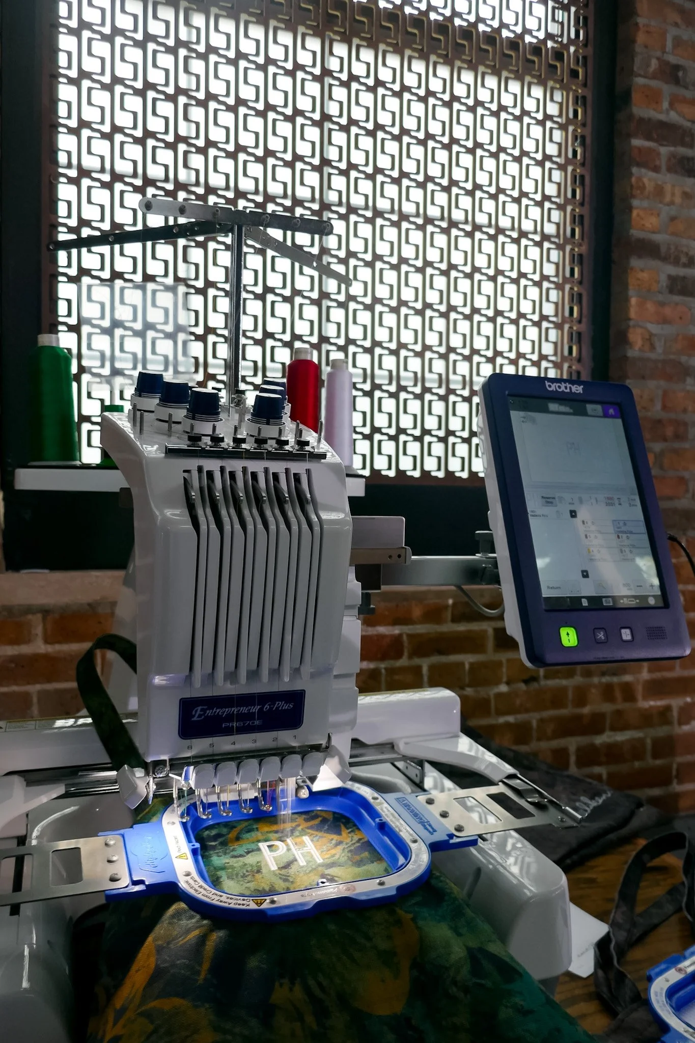 A sewing or embroidery machine with spools of thread in green, red, and white in the background, situated next to a Brother embroidery machine with a design reading 'PH' on the hoop. The setup is against a decorative brick wall with a geometric patterned window or partition.