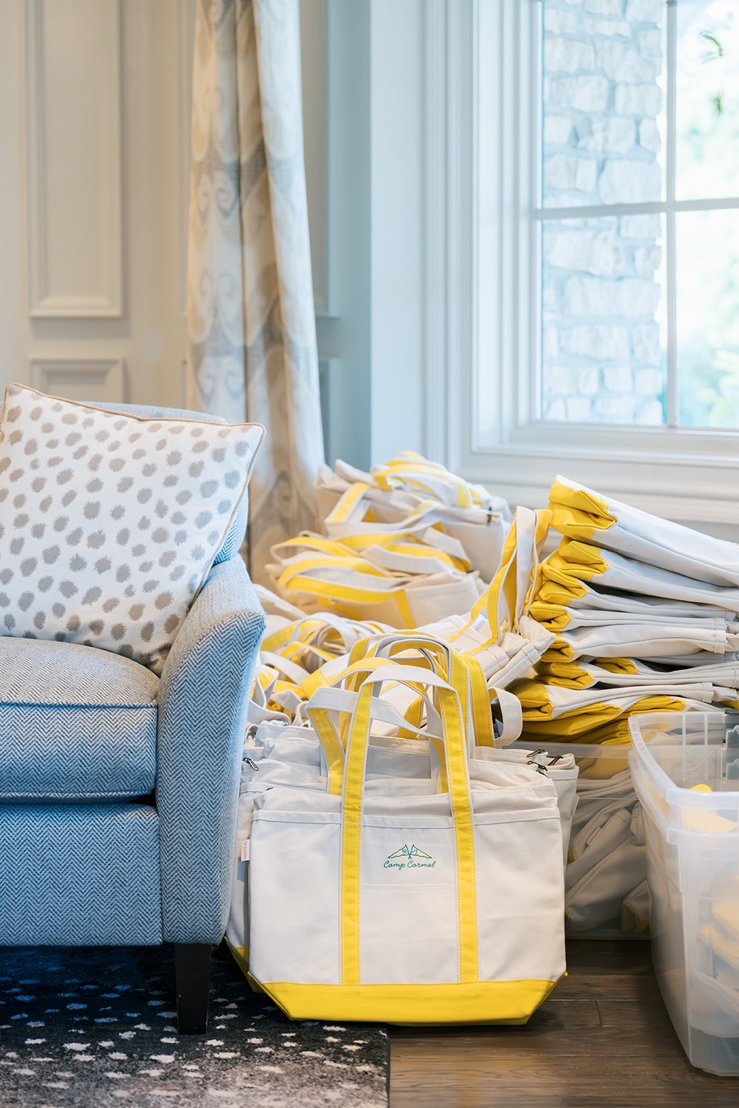Stacked white and yellow tote bags, yellow and white rolled and folded umbrellas or towels, and a beige armchair with a patterned pillow, next to a window with light curtains in a bright room.