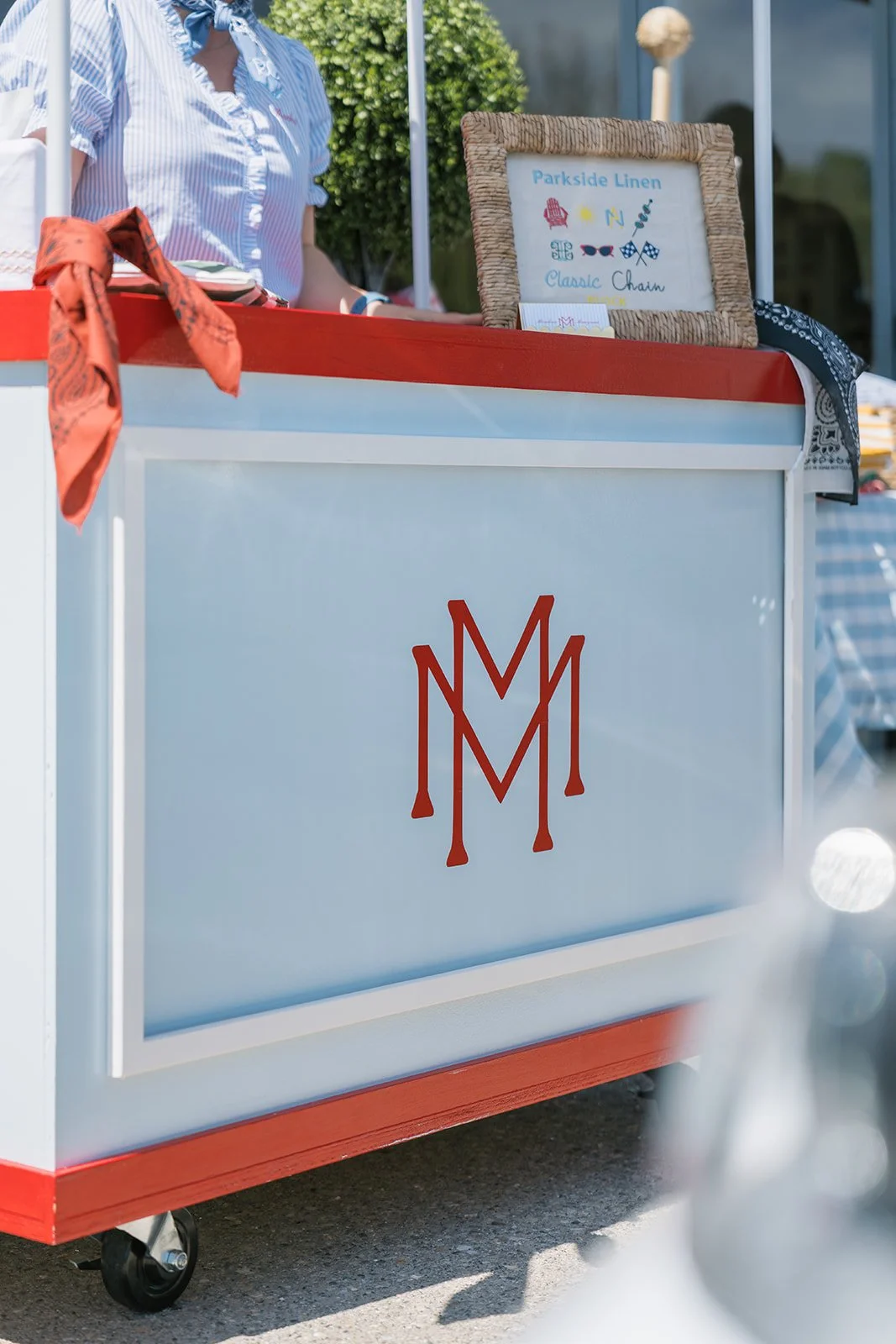 Side of a blue and red street cart with a monogram logo, selling linen products, at an outdoor market.