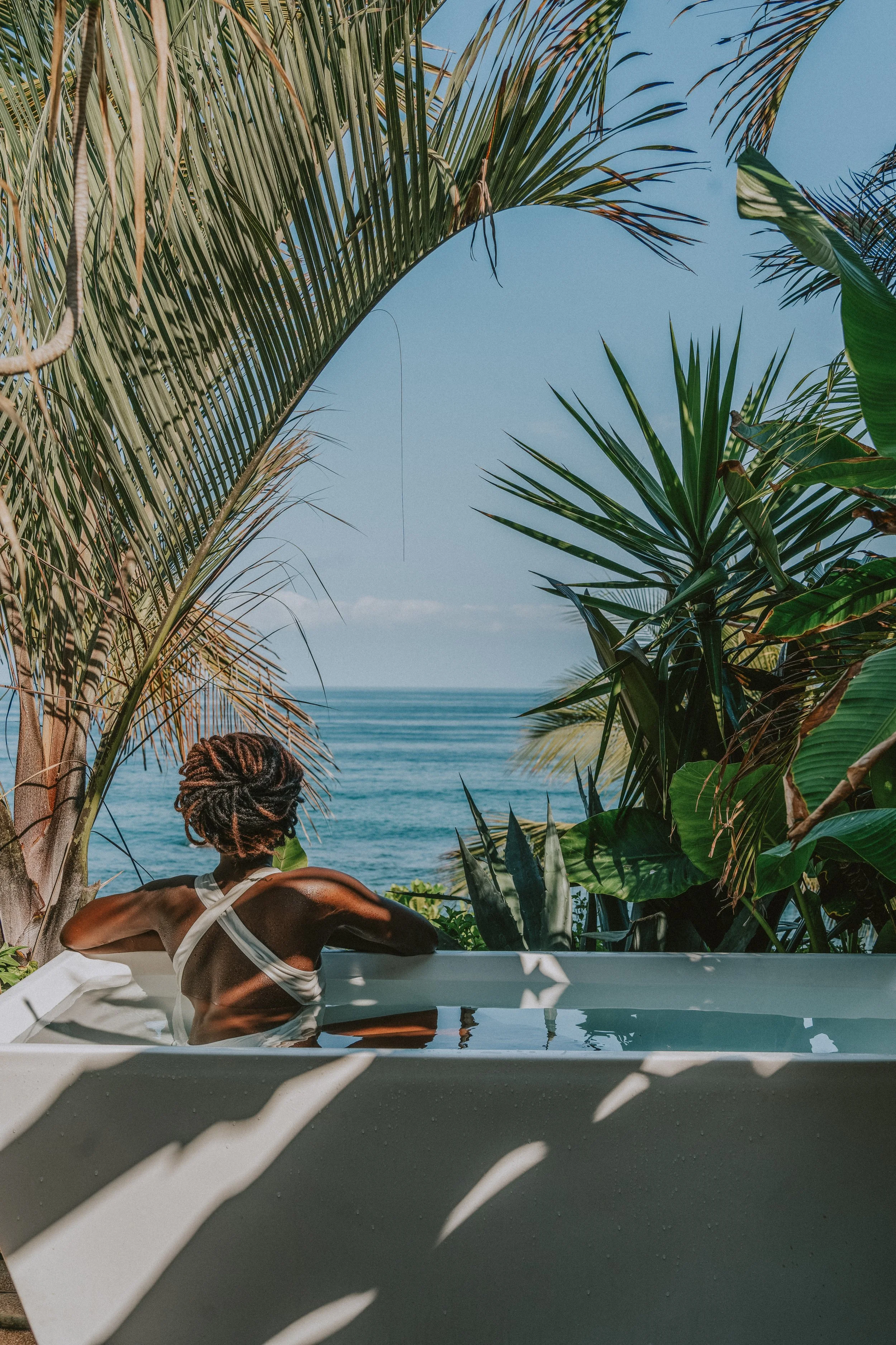 Une personne avec des dreadlocks relaxe dans une baignoire à côté de l'océan, entourée de végétation tropicale, sous un ciel ensoleillé. Photographe Assuama. www.assuama.co