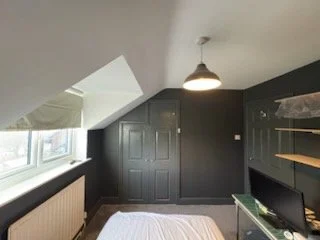 A bedroom with dark gray walls and built-in cabinets, a large window with a white curtain, and a bed with white linens. A ceiling light hangs from the white ceiling.