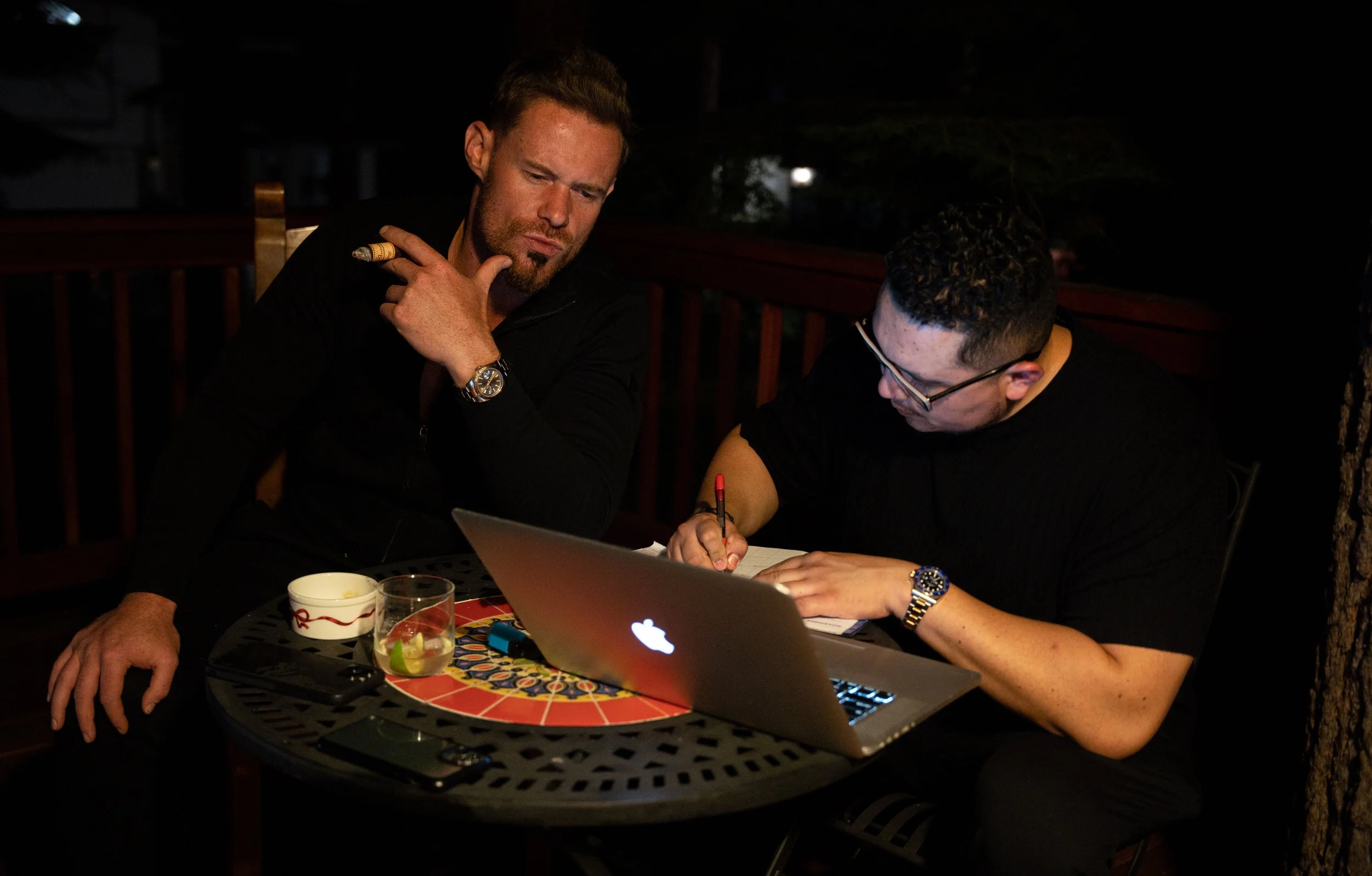 Two men sitting at a round outdoor table at night, working on a laptop and writing in a notebook, with drinks on the table.