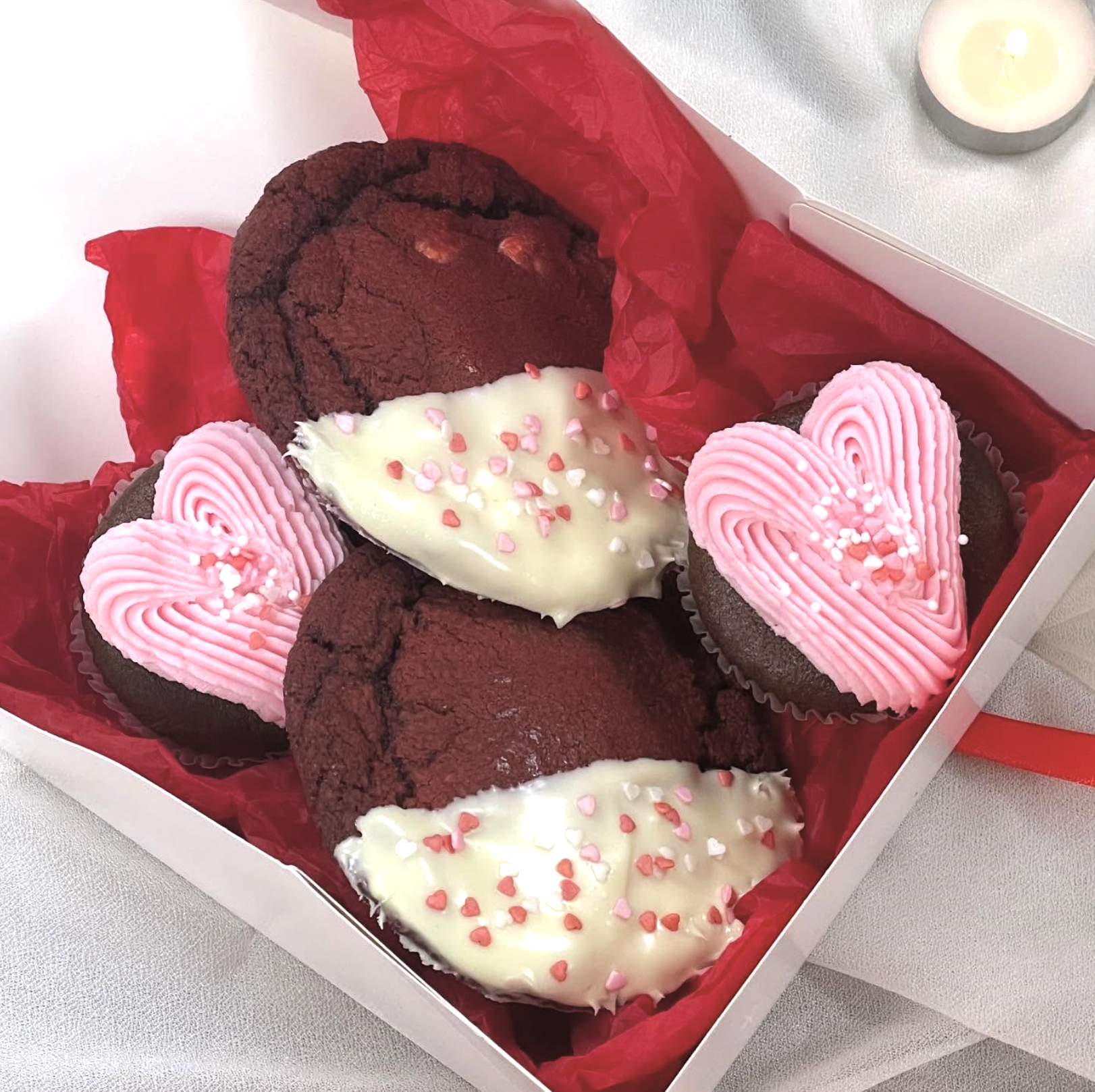 A box of Valentine's Day cookies with pink heart-shaped frosting, white hearts with pink and red sprinkles, and chocolate cookies with white chocolate hearts covered in pink and red sprinkles, all arranged on red tissue paper.