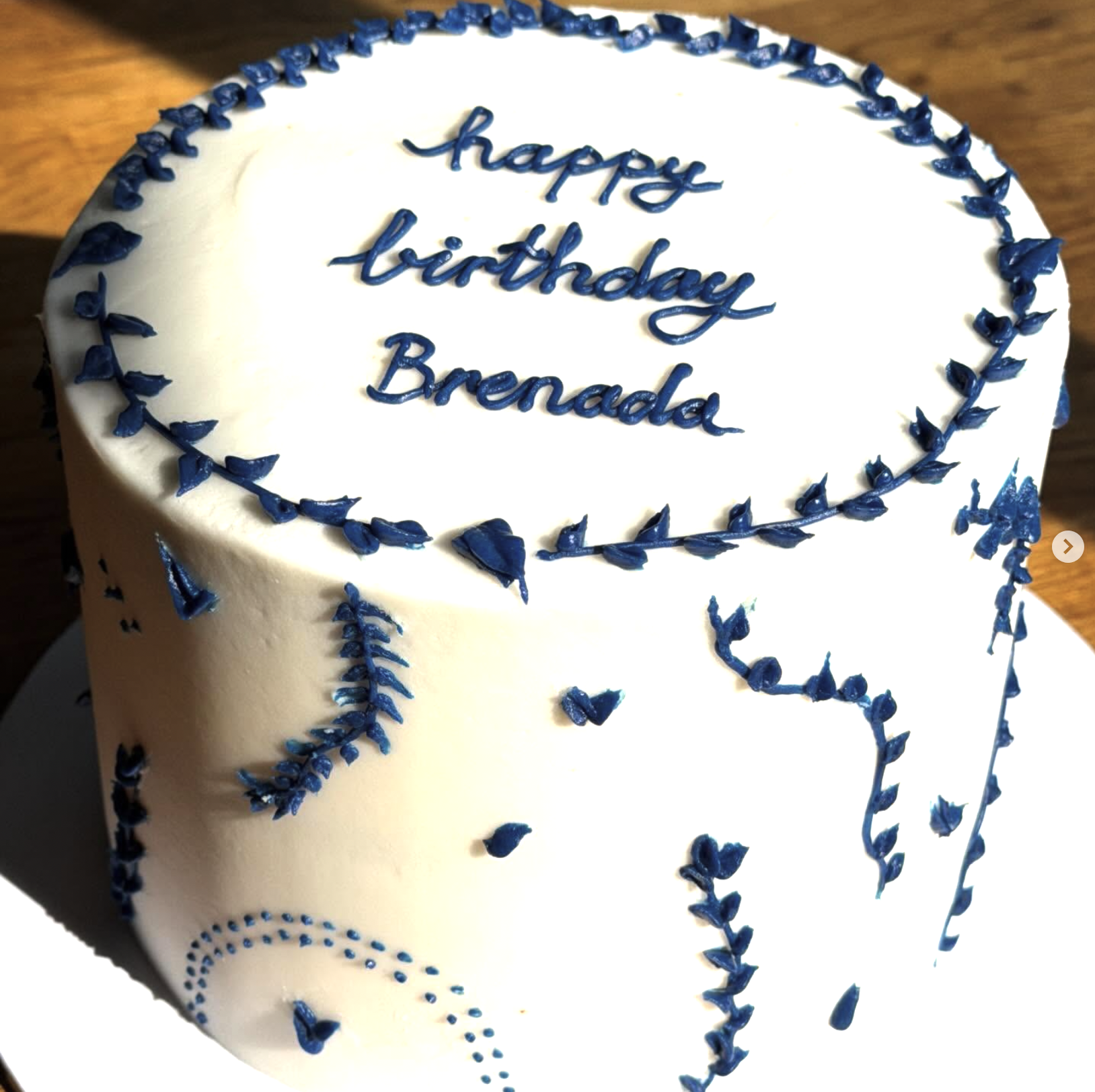 A white birthday cake decorated with blue icing, with the message 'happy birthday Brenda' written on top in blue cursive icing.