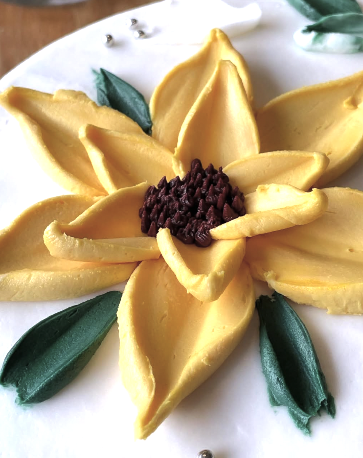 Decorative cake with yellow petals resembling a flower, green leaves, and a center made of dark chocolate chips.