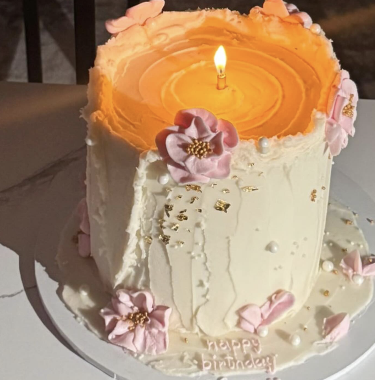 A birthday cake with a white frosting exterior decorated with pink flowers, white pearls, and gold accents. The cake has a single lit candle on top, which is surrounded by a peach-colored frosting that resembles a floating candle flame.