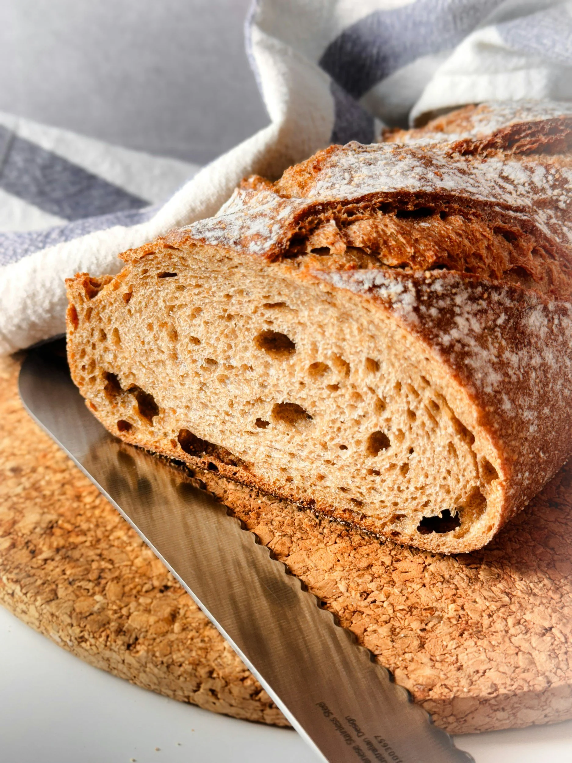 Sliced loaf of rustic bread with a crunchy crust, placed on a cork board with a serrated bread knife.