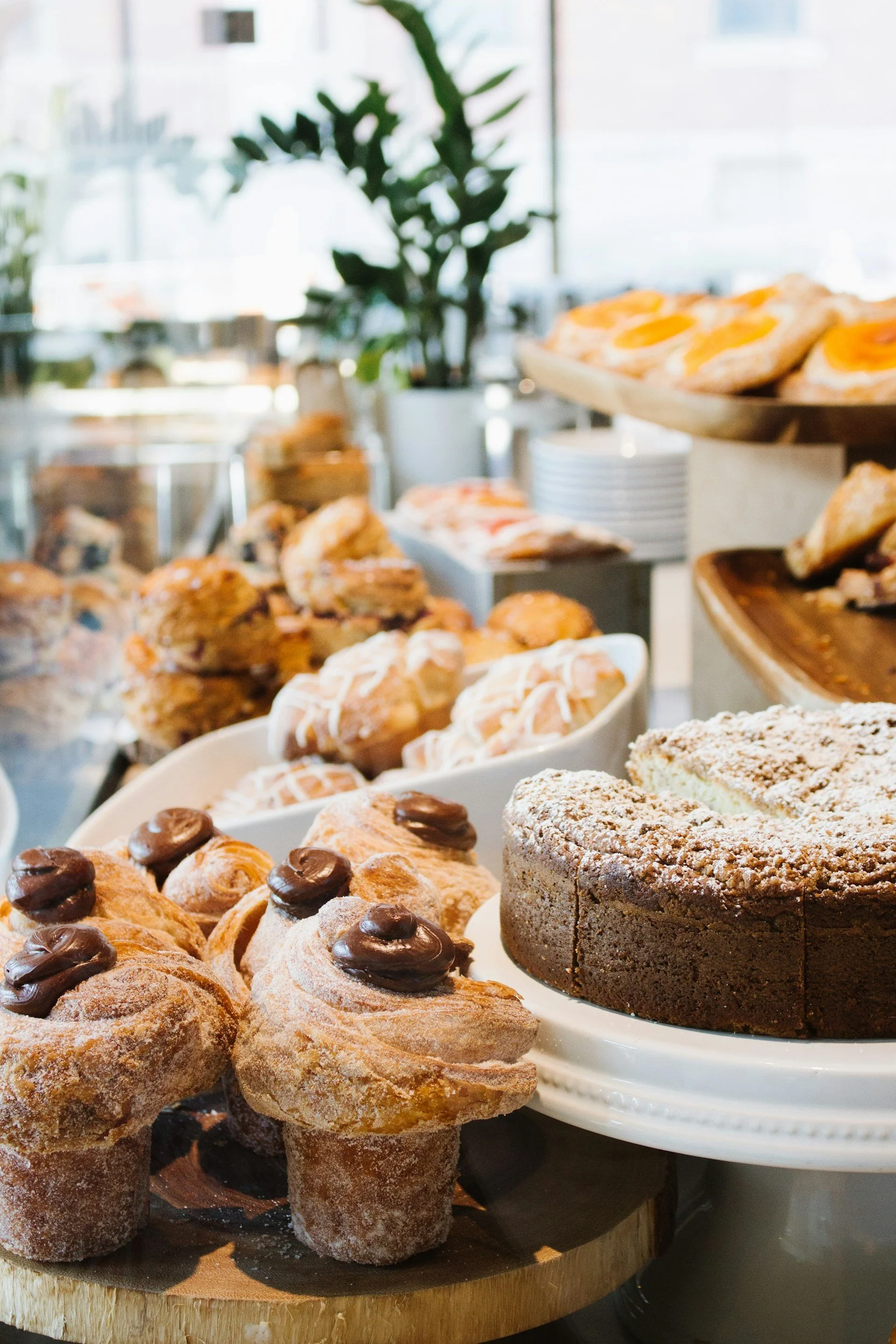 An assortment of baked goods including cinnamon rolls with chocolate frosting, layered cake, and scones, displayed on white plates and a wooden board in a brightly lit cafe.