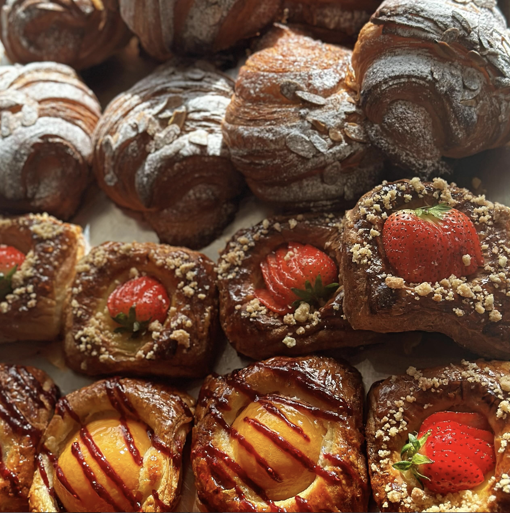 Assorted baked pastries including croissants dusted with powdered sugar, fruit tarts topped with strawberries and crumbles, and Danish pastries decorated with fresh strawberries and drizzled with glaze.