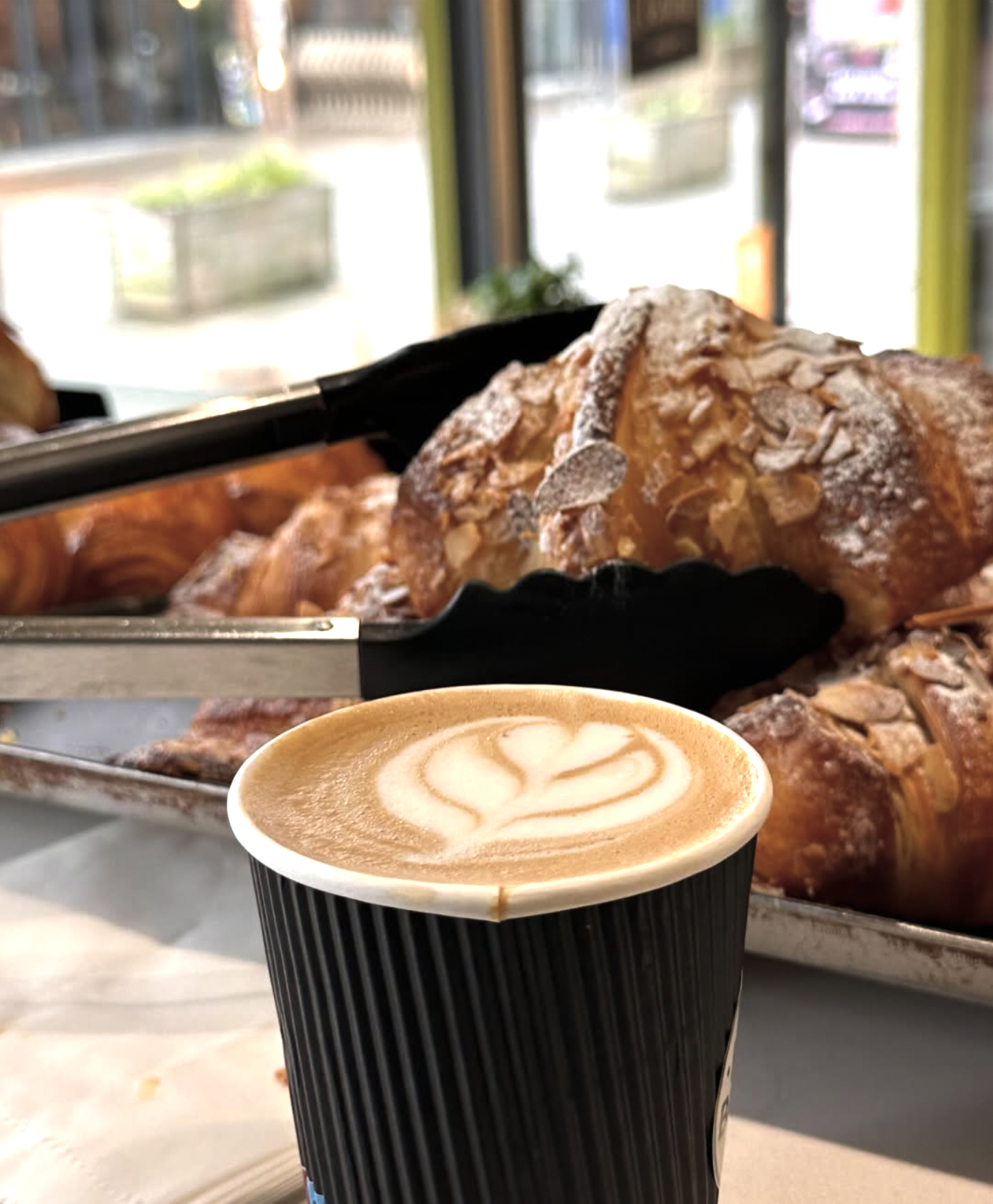 A hot latte with foam art in a black paper cup sits in front of a display of croissants with powdered sugar on top at a bakery or cafe.