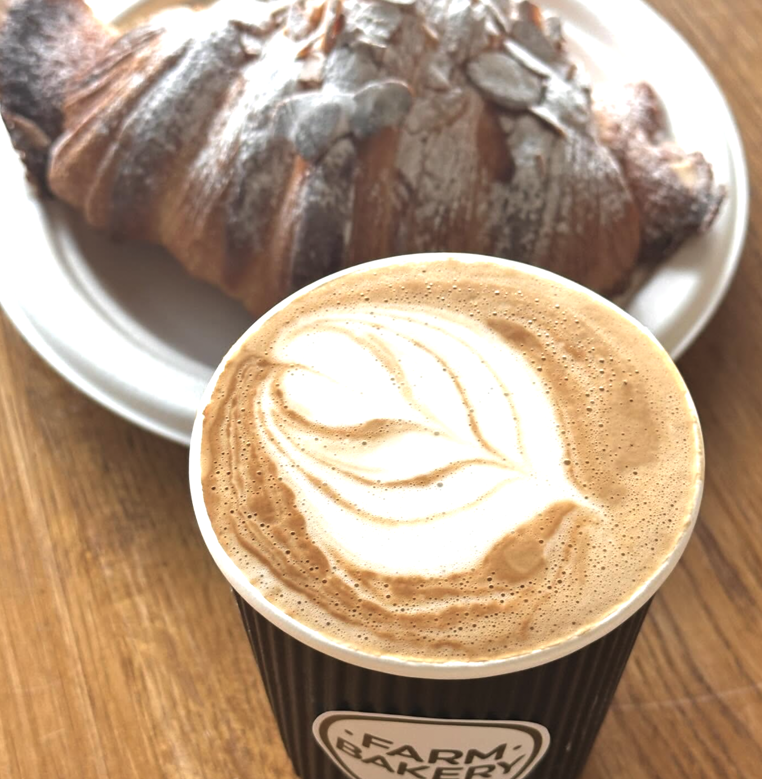 A cup of coffee with latte art on top and a croissant on a white plate in the background, placed on a wooden surface.