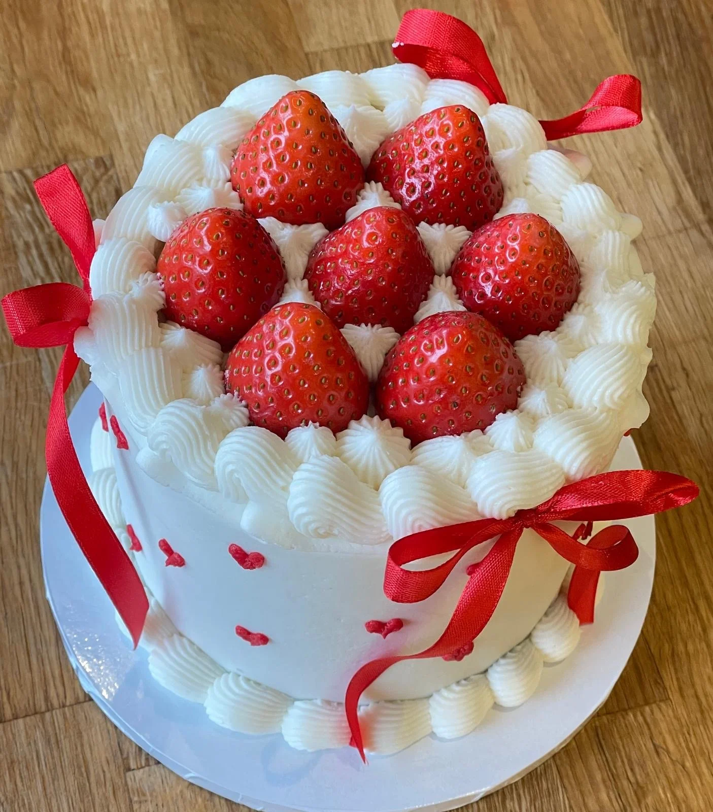 A round white cake decorated with fresh strawberries, white whipped cream, and red ribbons on a wooden surface.