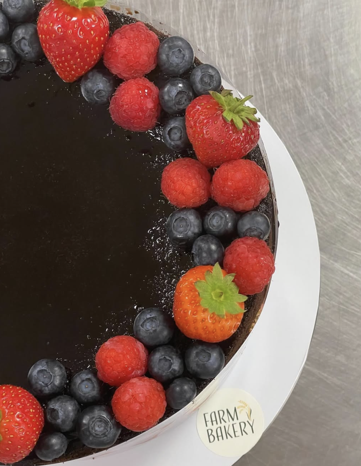 A round cake with a glossy dark chocolate glaze, decorated with fresh strawberries, raspberries, and blueberries on top.