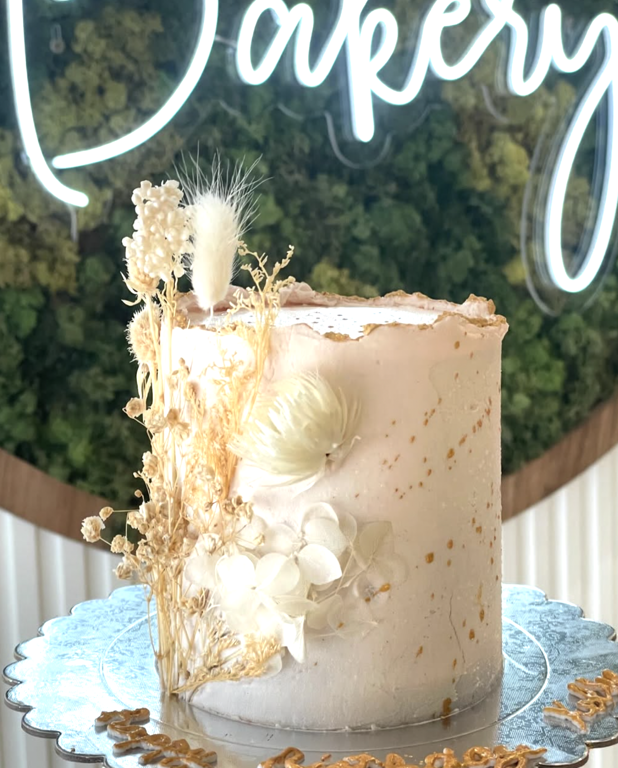 A tall white frosted cake decorated with dried flowers and a feather, with a backdrop of a neon sign and greenery.