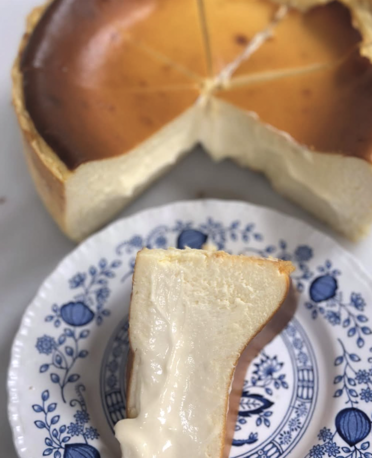 A slice of creamy cheesecake on a blue and white floral-patterned plate with a larger whole cheesecake in the background.
