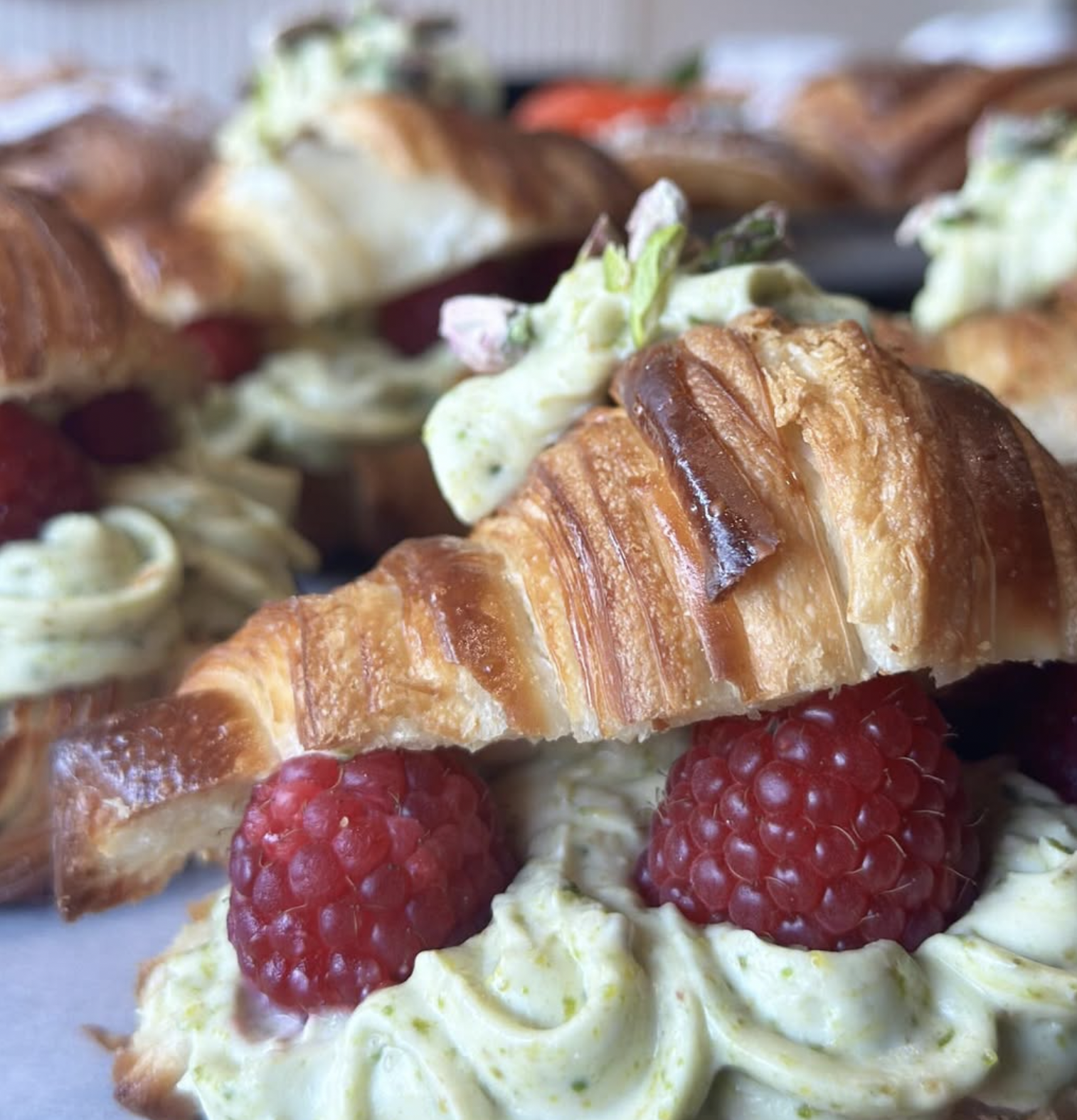 Close-up of a pastry topped with raspberries, whipped cream, and small pink flowers, with a blurred background of more pastries.