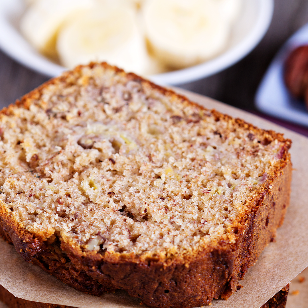 A slice of banana nut bread on parchment paper with a bowl of sliced bananas in the background.