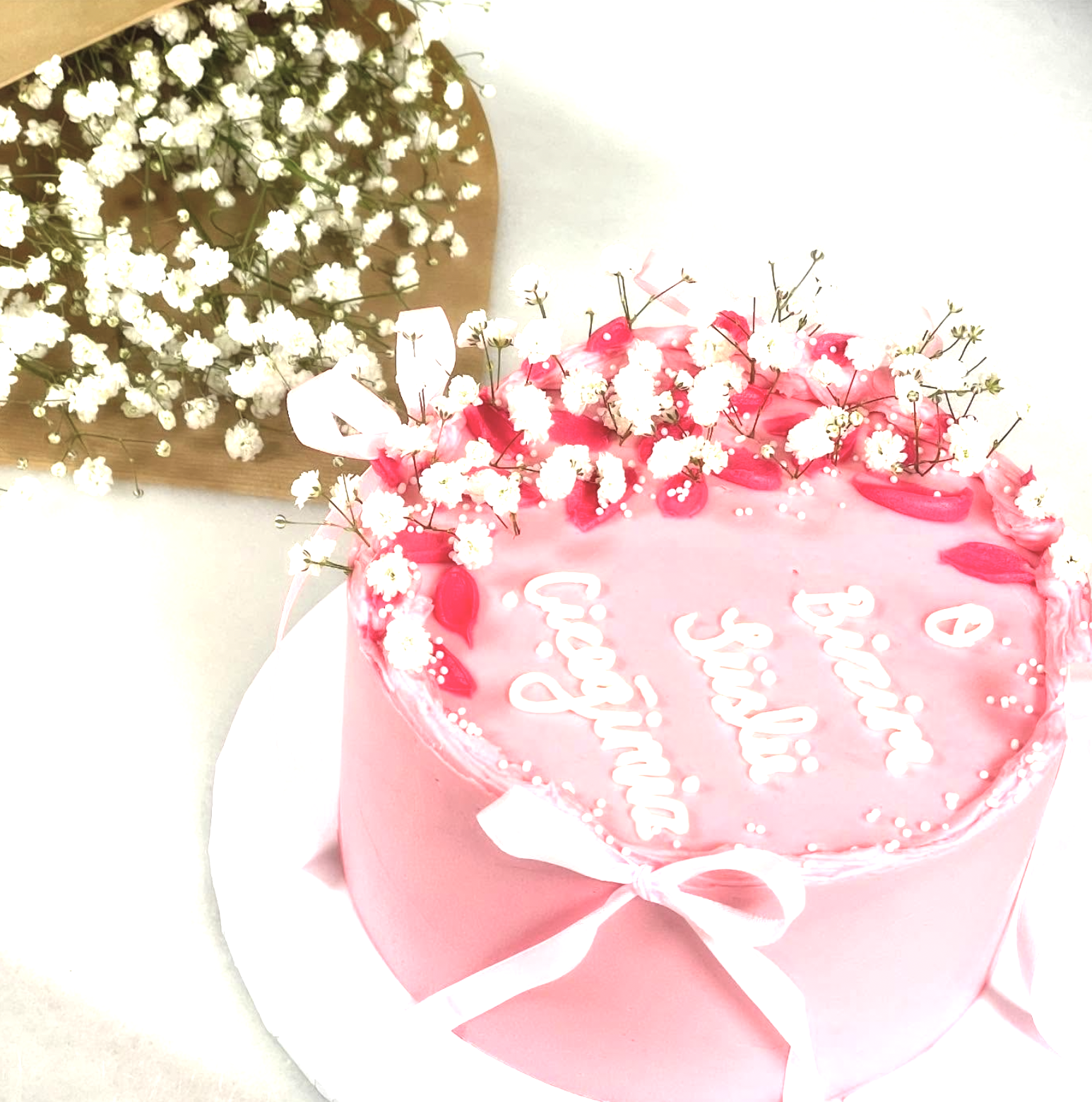 Pink birthday cake decorated with whipped cream and pink icing, adorned with small white flowers and pink petal-like decorations, with the message "Happy Birthday" written on top, set on a white surface. A bouquet of white baby's breath flowers is in the background.