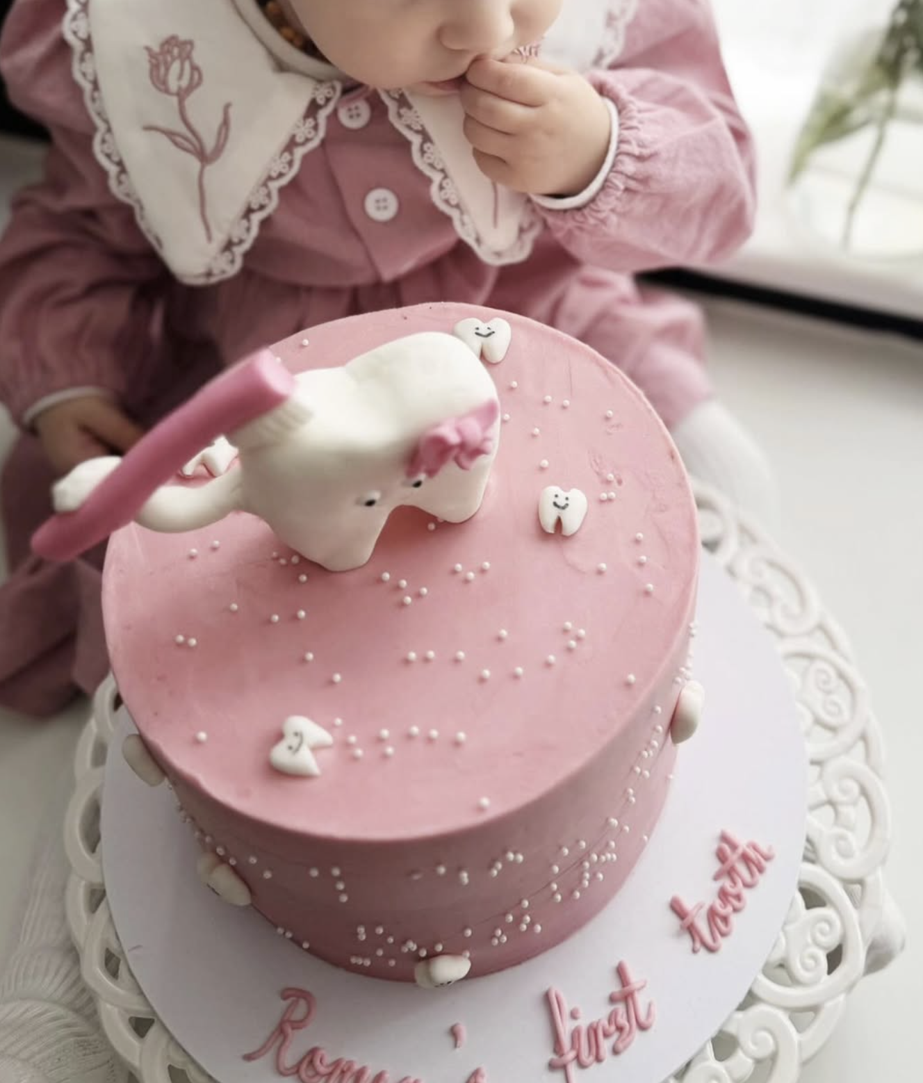 A young child in pink clothing and a embroidered white bib is sitting near a pink birthday cake. The cake has a unicorn figure on top and small tooth-shaped decorations with smiley faces, along with tiny white beads. The cake is on a white decorative