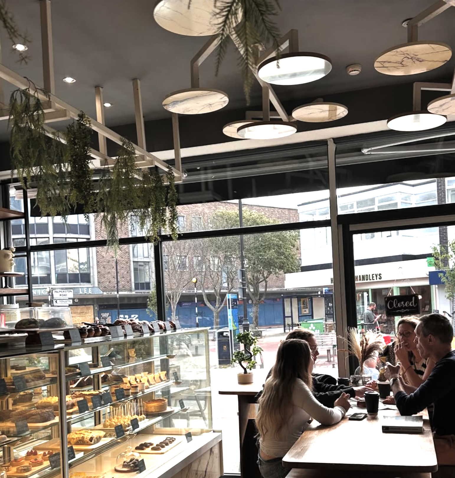 A bakery cafe with a glass display case filled with various pastries and cakes. Inside, four people are sitting at a wooden table near the window, engaged in conversation. The cafe has modern lighting fixtures on the ceiling and hanging greenery. Out