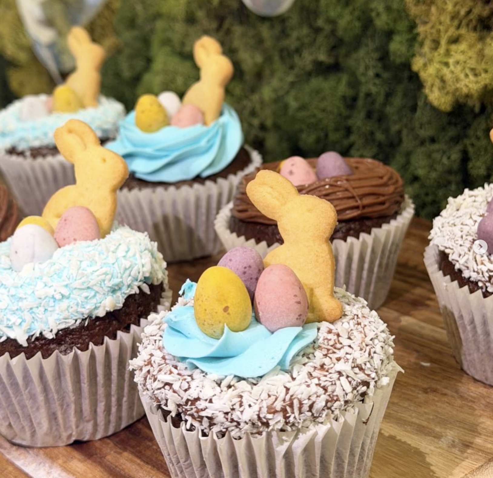 Several decorated cupcakes with Easter themed toppings, including bunny-shaped cookies, colorful candy eggs, and pastel-colored frosting, displayed on a wooden surface.