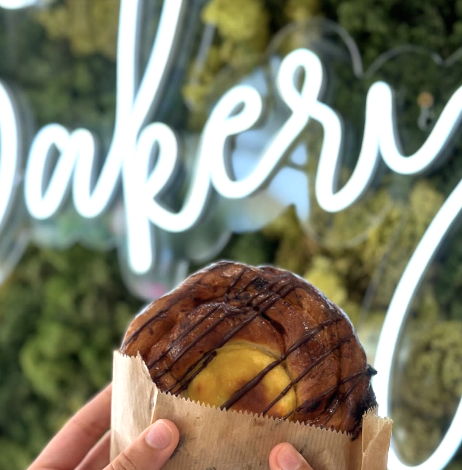 A person holding a baked pastry with a yellow filling, drizzled with chocolate, and in a brown paper bag. In the background, there is a neon sign that spells out 'cafe' and some greenery.