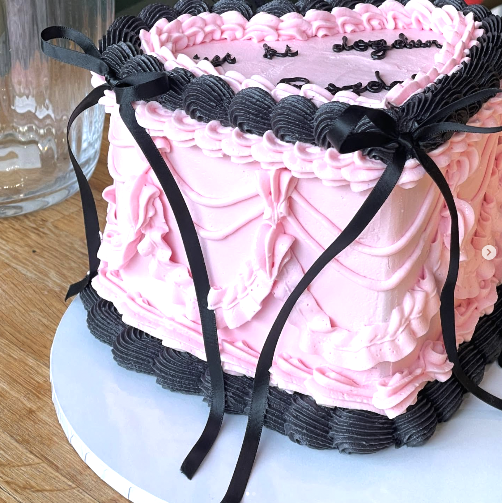 Pink and black heart-shaped birthday cake with piped frosting, black ribbon decorations, and writing on top, placed on a silver cake board.