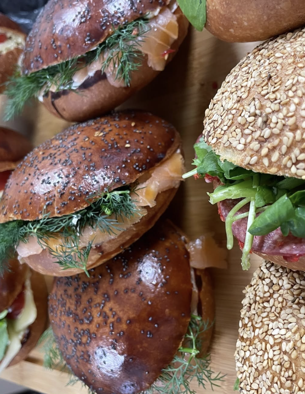 Close-up of assorted mini sandwiches on a wooden platter, featuring poppy seed and sesame seed buns with various fillings like smoked salmon, greens, and deli meat.