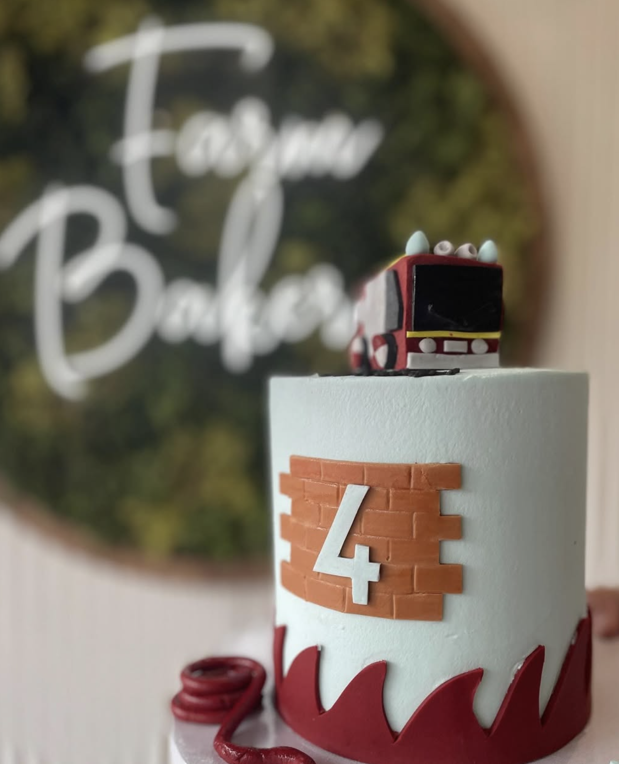 A small cake decorated with a brick pattern and the number 4, topped with a small red bus toy, with a mirror in the background reflecting parts of the cake and the words 'treat & balace'.